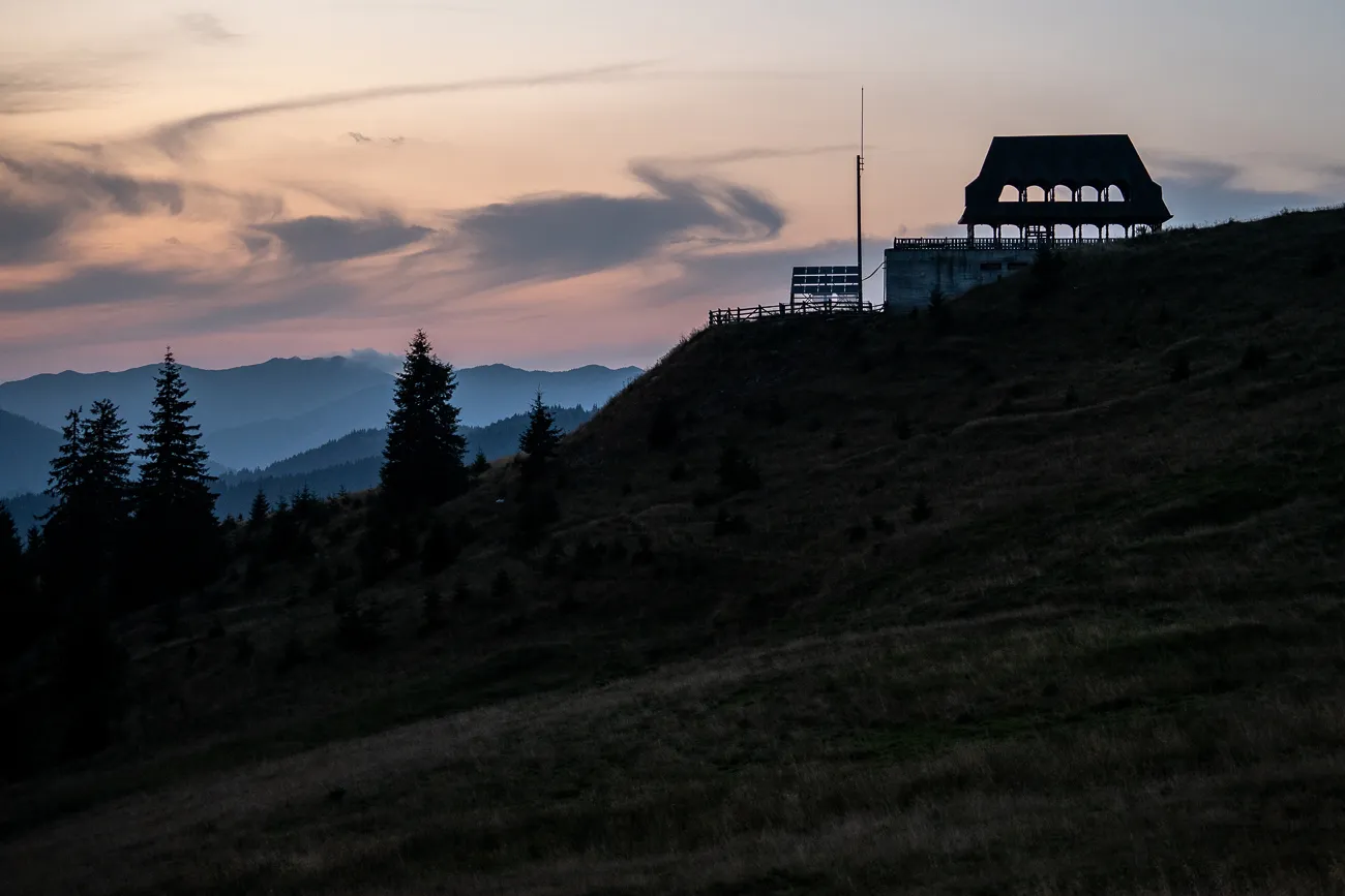 Silhouette einer Aussichtsplattform auf einem Hügel bei Sonnenuntergang mit Bergen im Hintergrund.