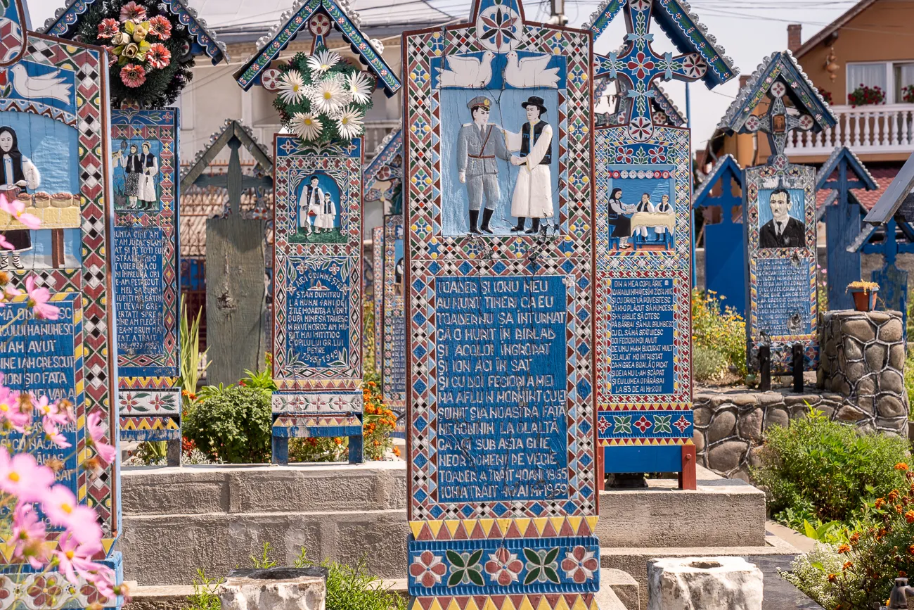 Farbenfrohe, bemalte Holzgrabkreuze in einem Friedhof mit Blumen und Grünzeug.