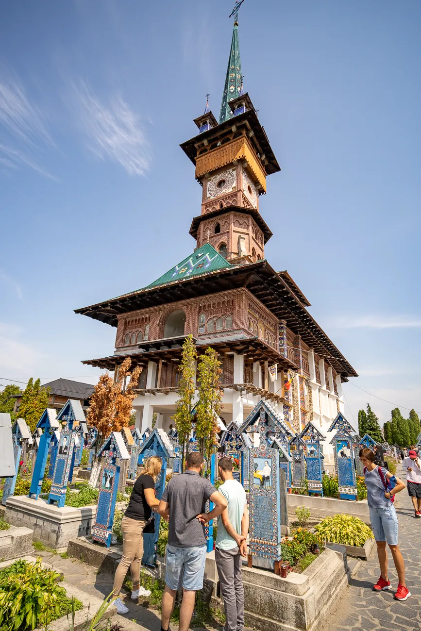 Besucher betrachten farbenfrohe Grabkreuze vor einer Holzkirche mit Turm.