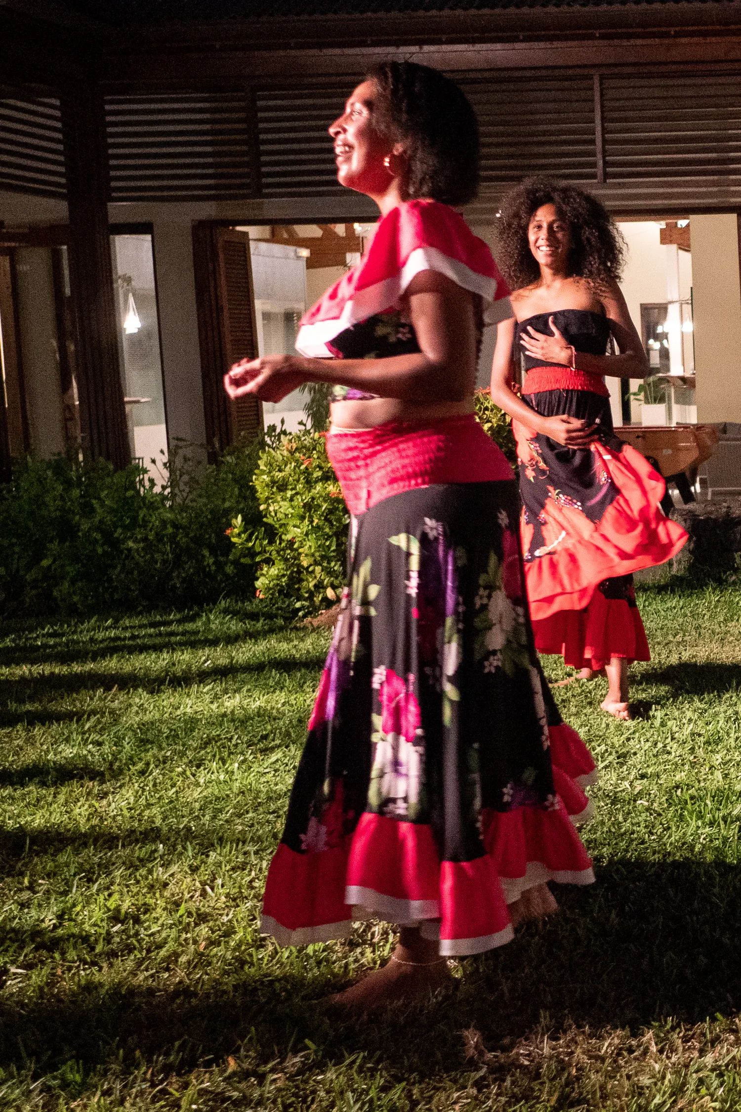 Zwei Frauen tanzen im Garten bei Nacht, bekleidet mit farbenfrohen traditionellen Kleidern.