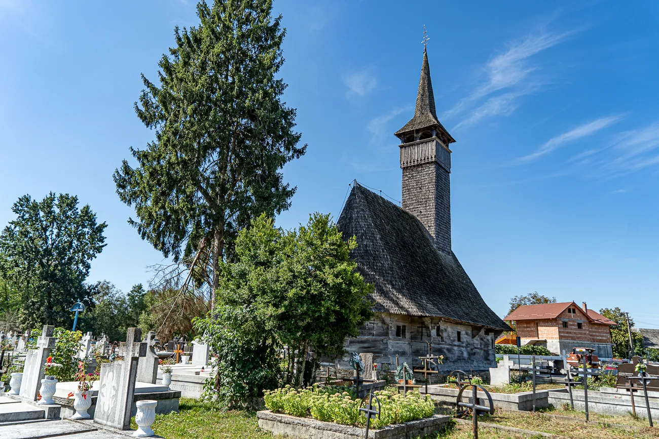 Holzbau mit Turm und Dach, umgeben von Bäumen und Grabsteinen unter blauem Himmel
