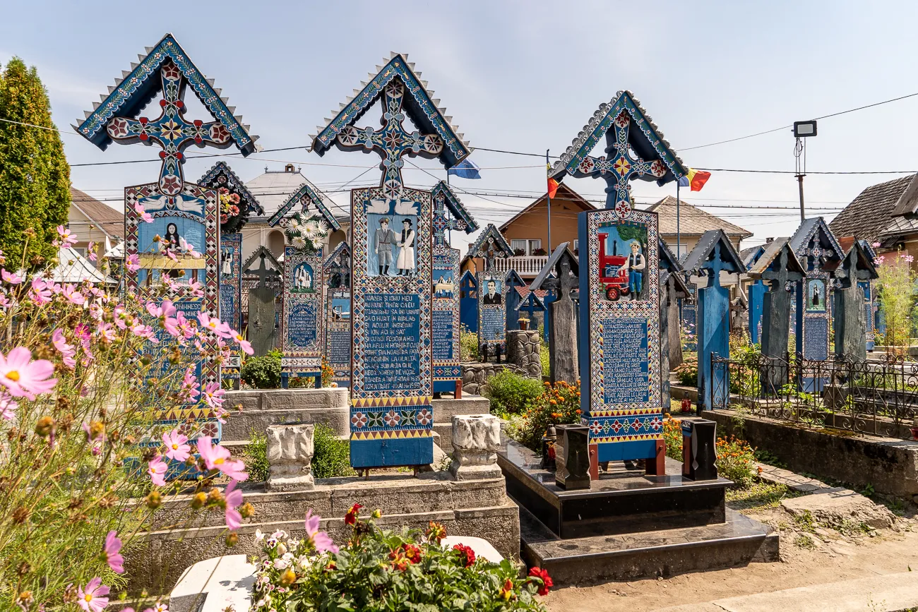 Farbige Holzkreuze mit Bildern und Texten in einem Friedhof, umgeben von Blumen.