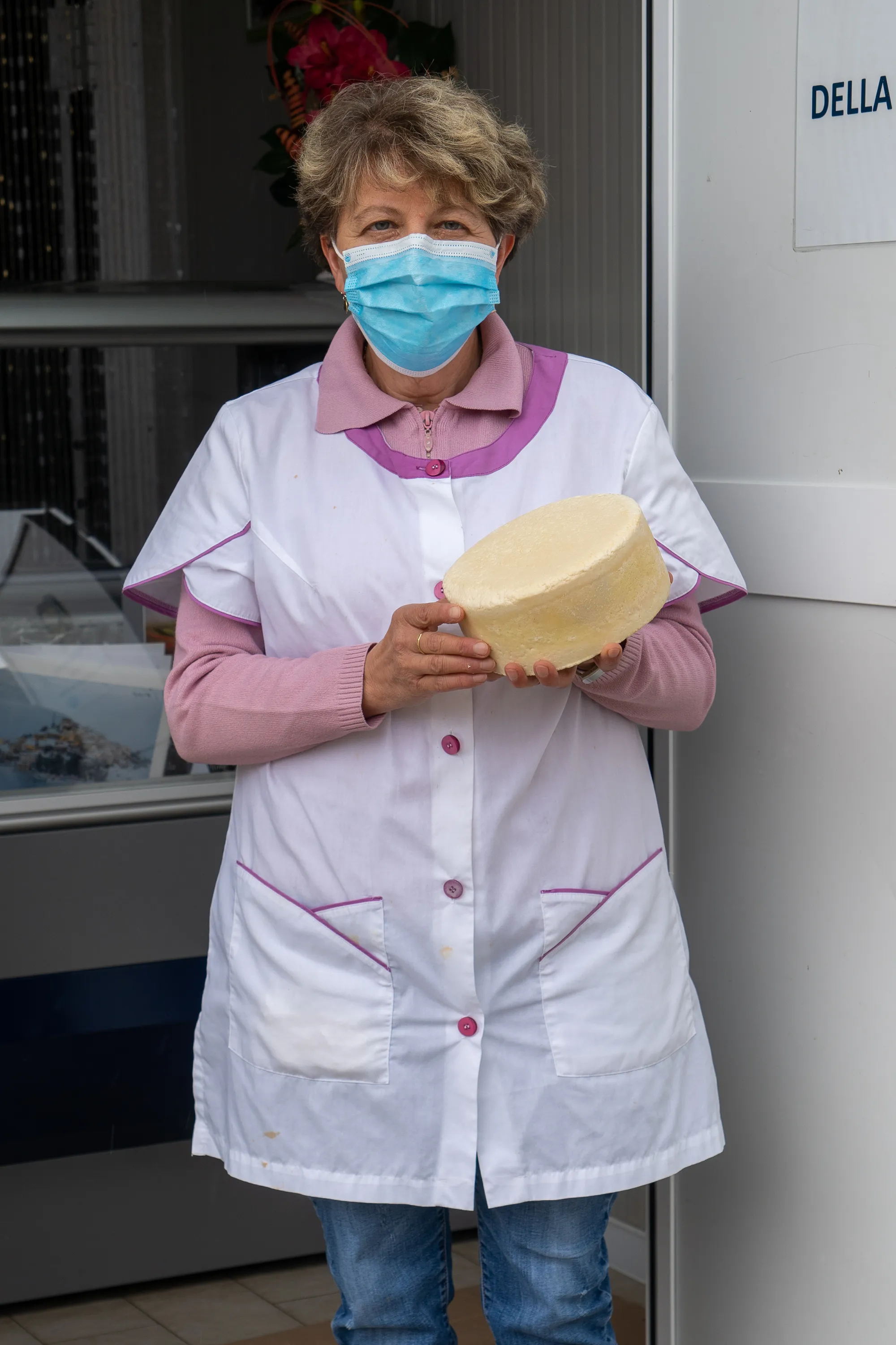 Frau in weißer Schürze hält runden Käse vor Geschäftsfassade