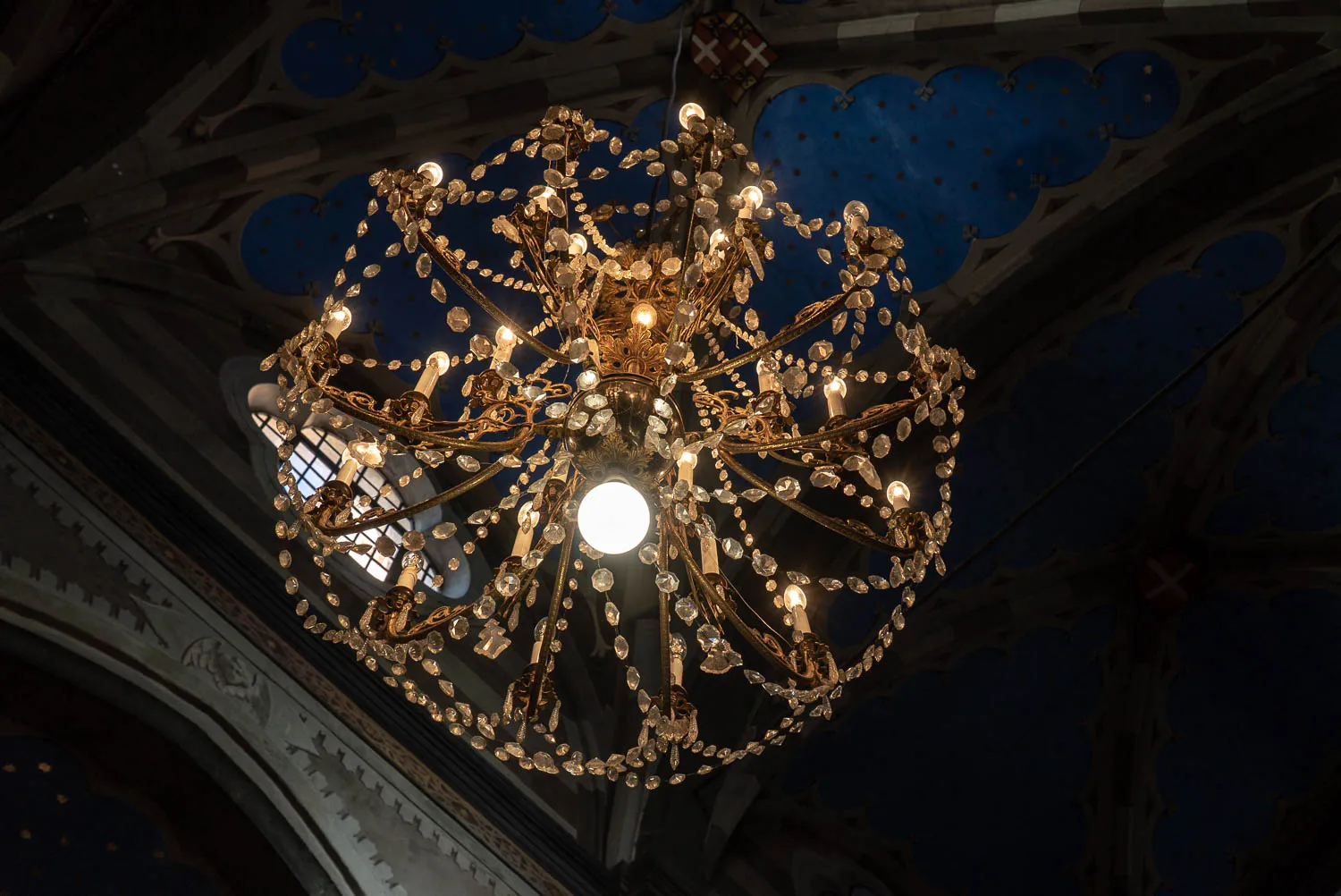 Goldschmiede Kronleuchter mit Kristallen unter blau bemaltem Gewölbe.