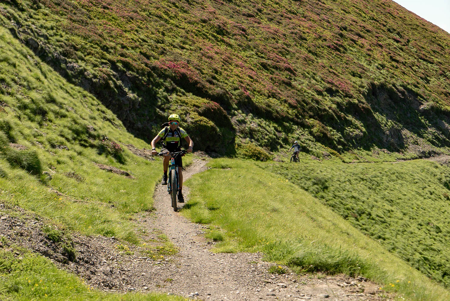 Zwei Mountainbiker fahren auf einem steilen grasbewachsenen Pfad durch einen Hügel.