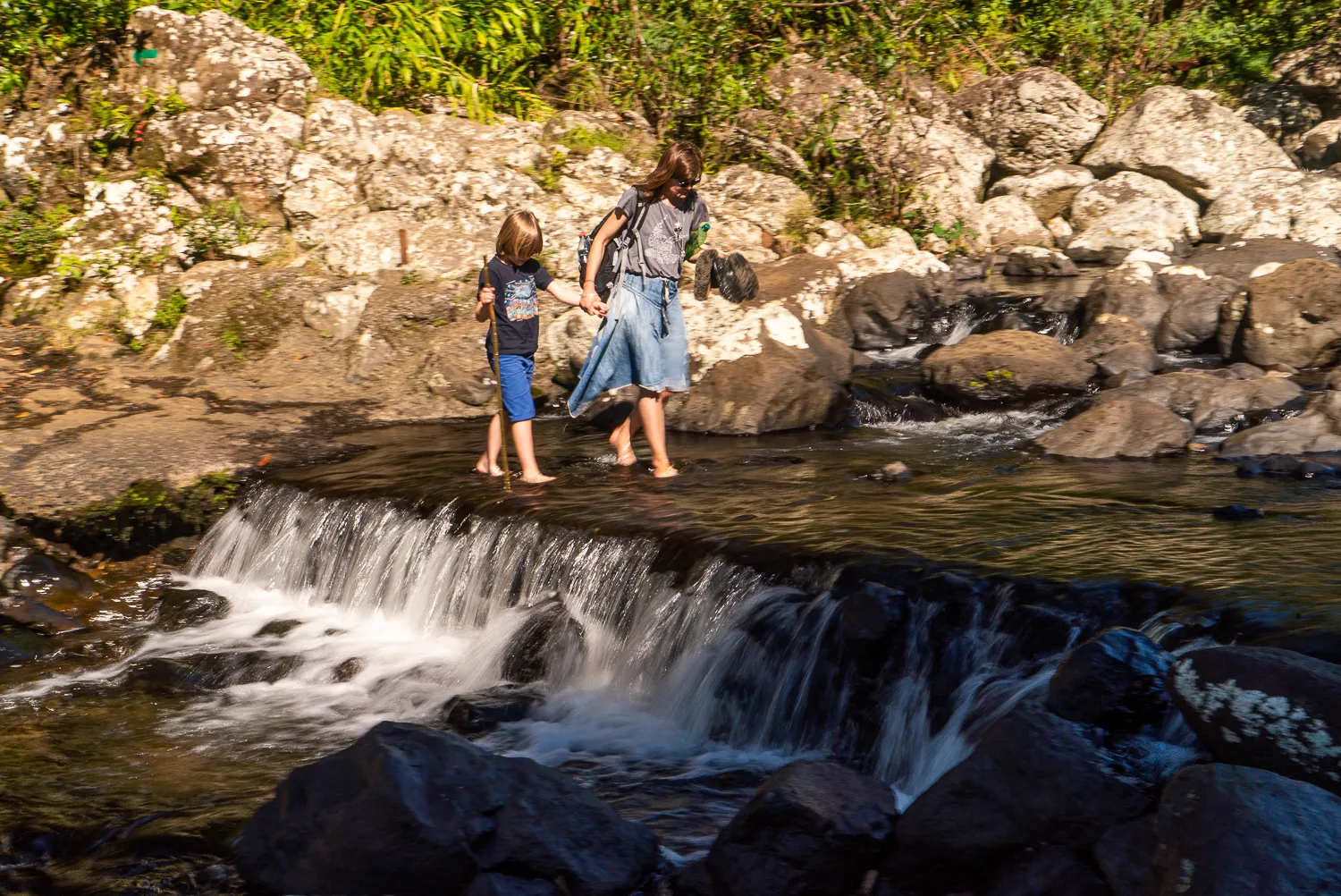 Zwei Personen gehen barfuß über einen kleinen Wasserfall in einem steinigen Flussbett.