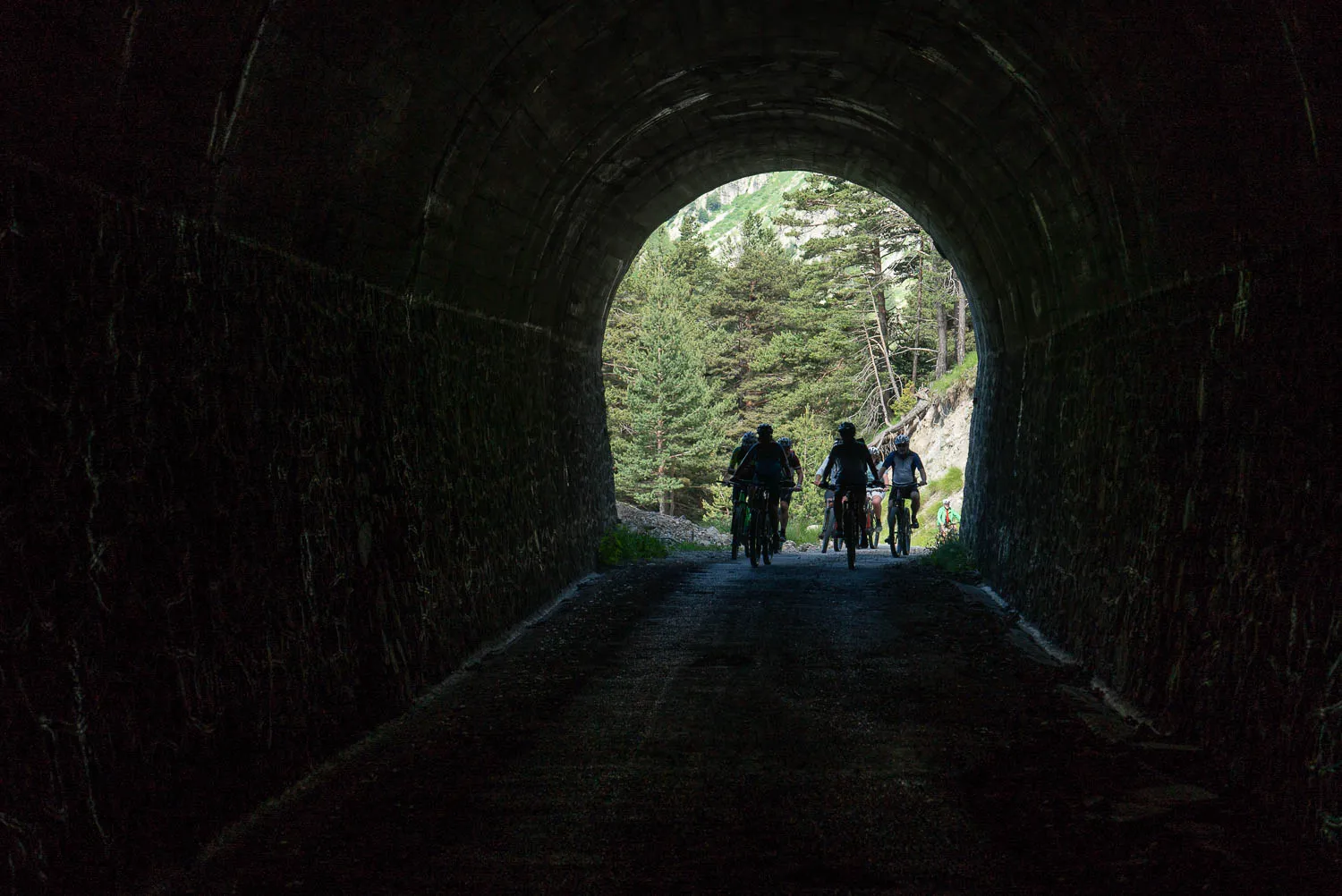 Drei Radfahrer fahren durch einen dunklen Steinbogen in den Wald hinaus.