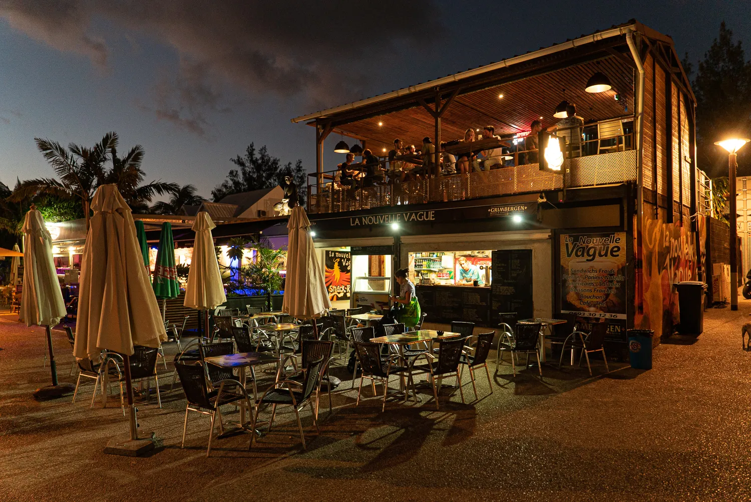 Beleuchteter Außenbereich eines Restaurants mit Tischen, Stühlen und geschlossenen Sonnenschirmen bei Nacht.