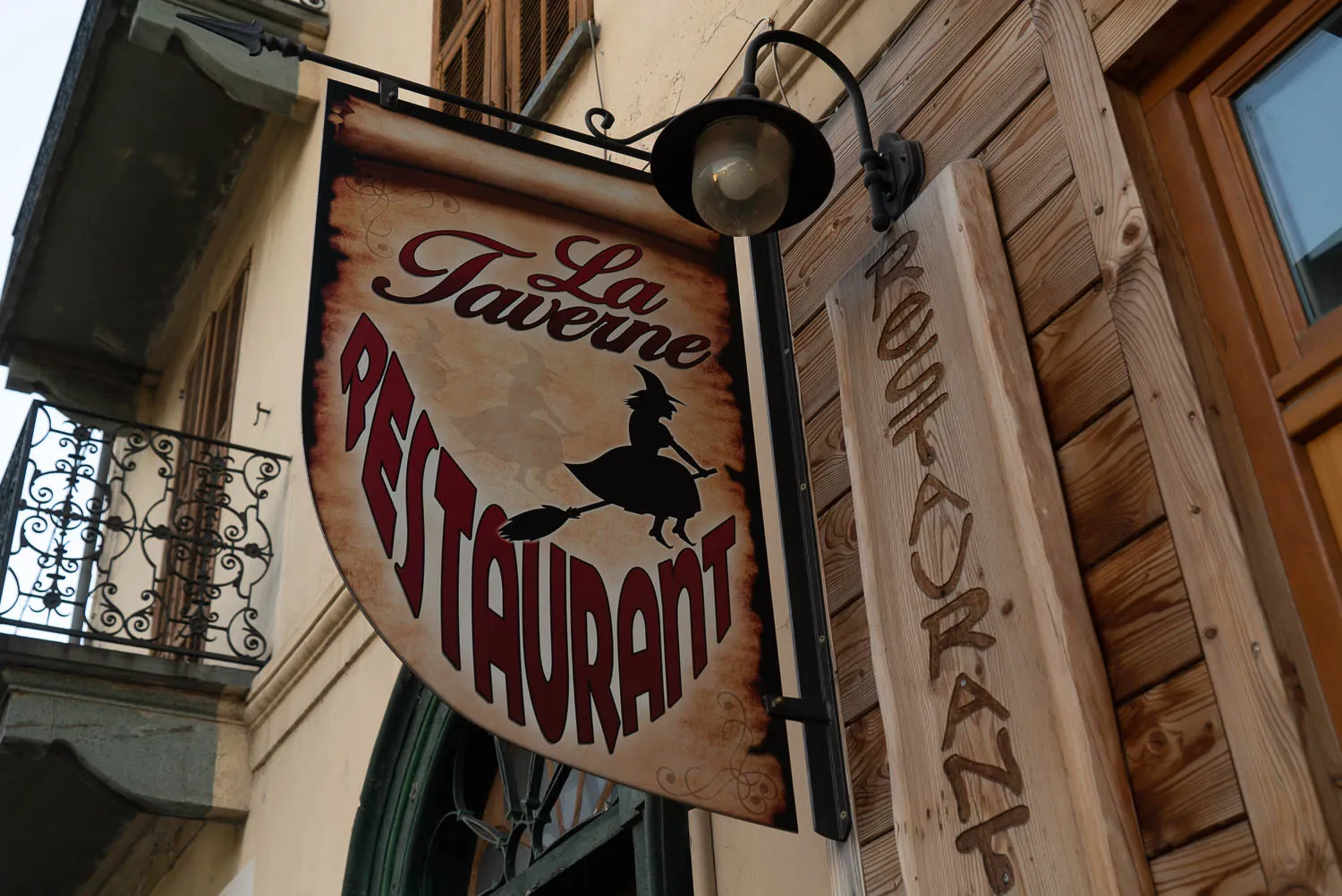 Schild eines Restaurants mit Hexe auf Besen, an Holzwand und Gebäude angebracht
