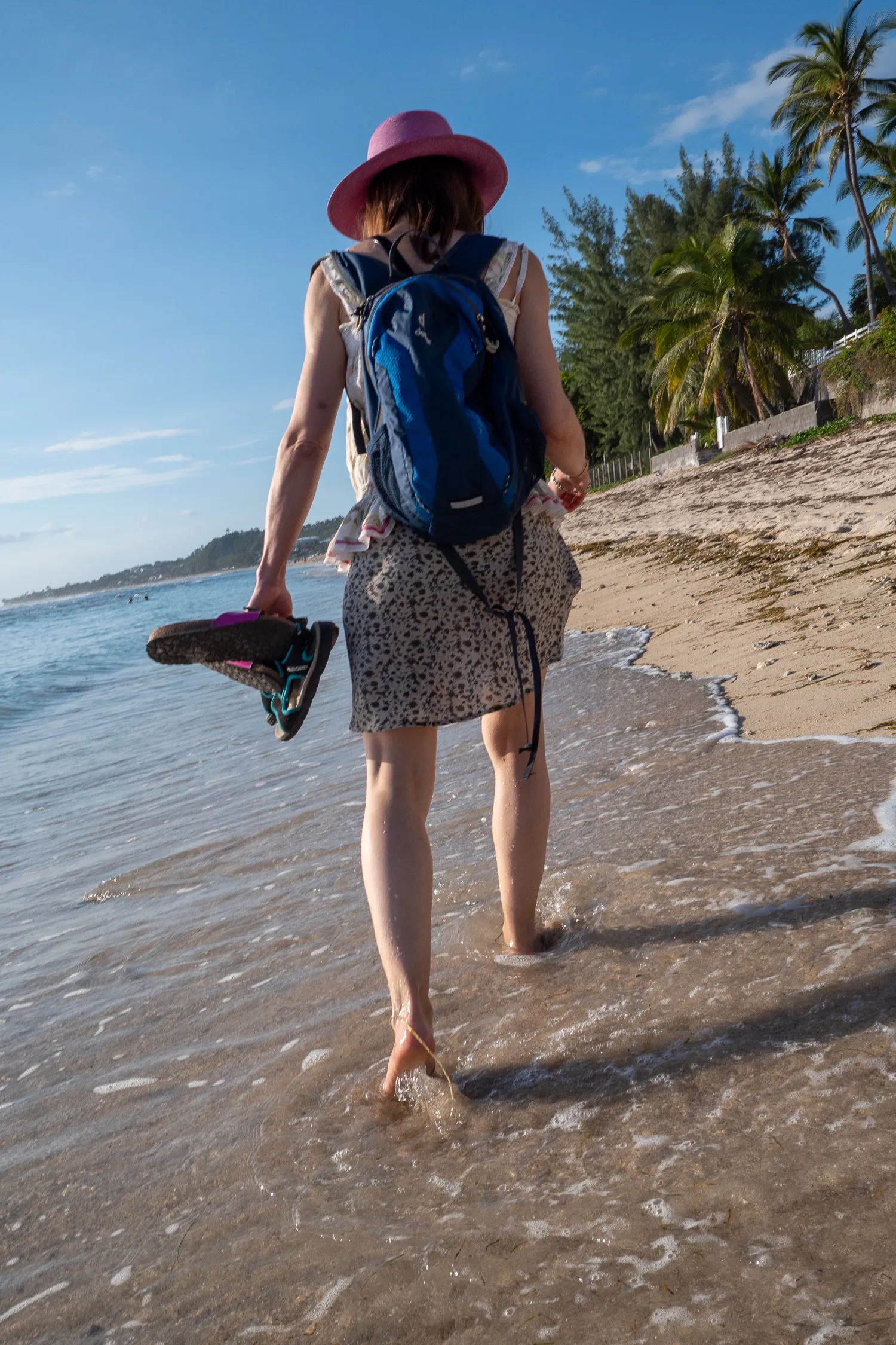 Person mit Rucksack und Schuhen am Strand im flachen Wasser