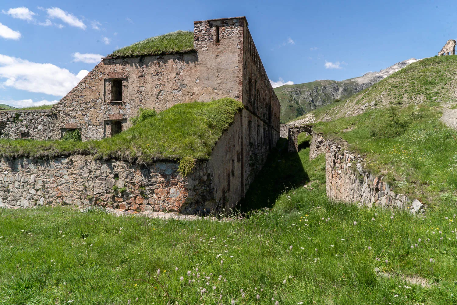 Ruine eines Steinbaus mit Grasdach in grüner Berglandschaft unter blauem Himmel.