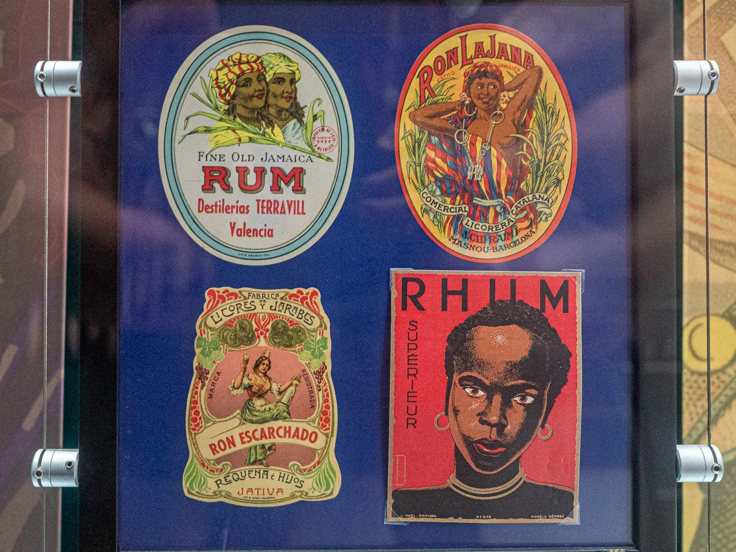 Vier historische Rumetiketten in einem Glasdisplay mit blauem Hintergrund.
