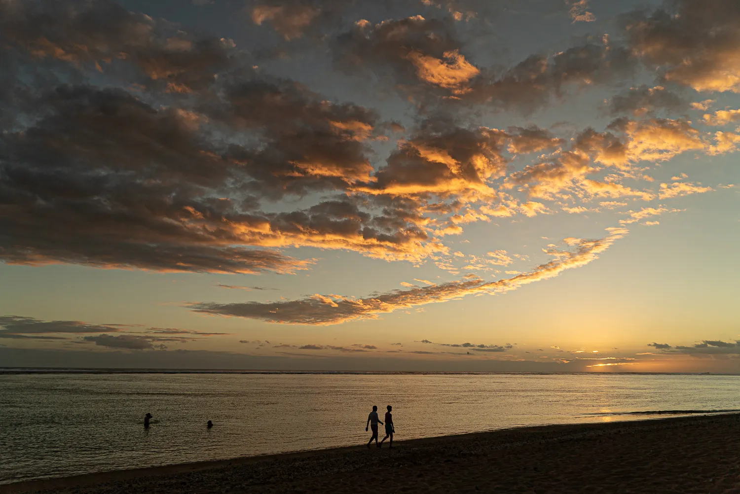 Zwei Silhouetten gehen am Strand entlang bei Sonnenuntergang mit orangefarbenen Wolken.