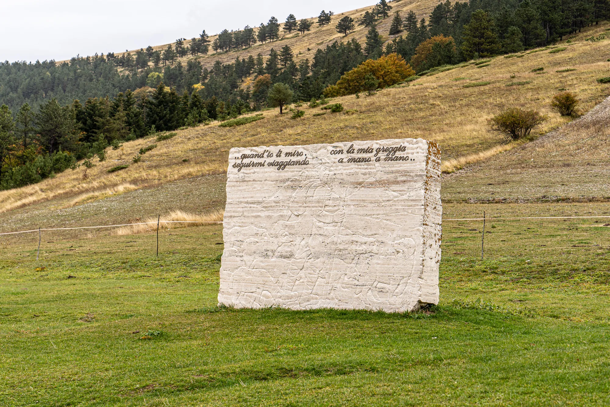 großer weißer Stein mit eingraviertem Text in einer grünen Wiese vor bewaldeten Hügeln