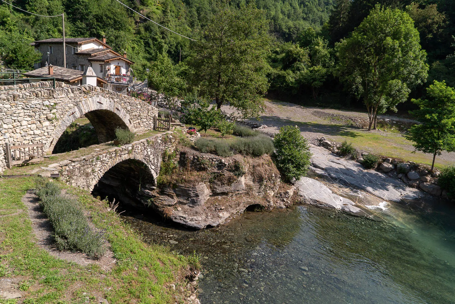 Steinbrücke über Fluss mit Ufervegetation und Häusern im Hintergrund