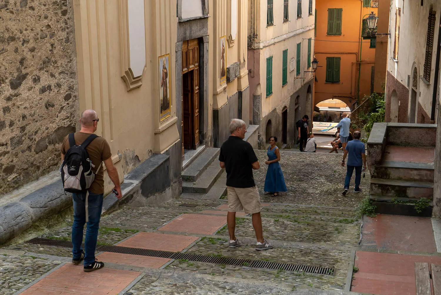 Menschen gehen eine steinerne Gasse entlang, Gebäude mit Fenstern und Türen im Hintergrund.