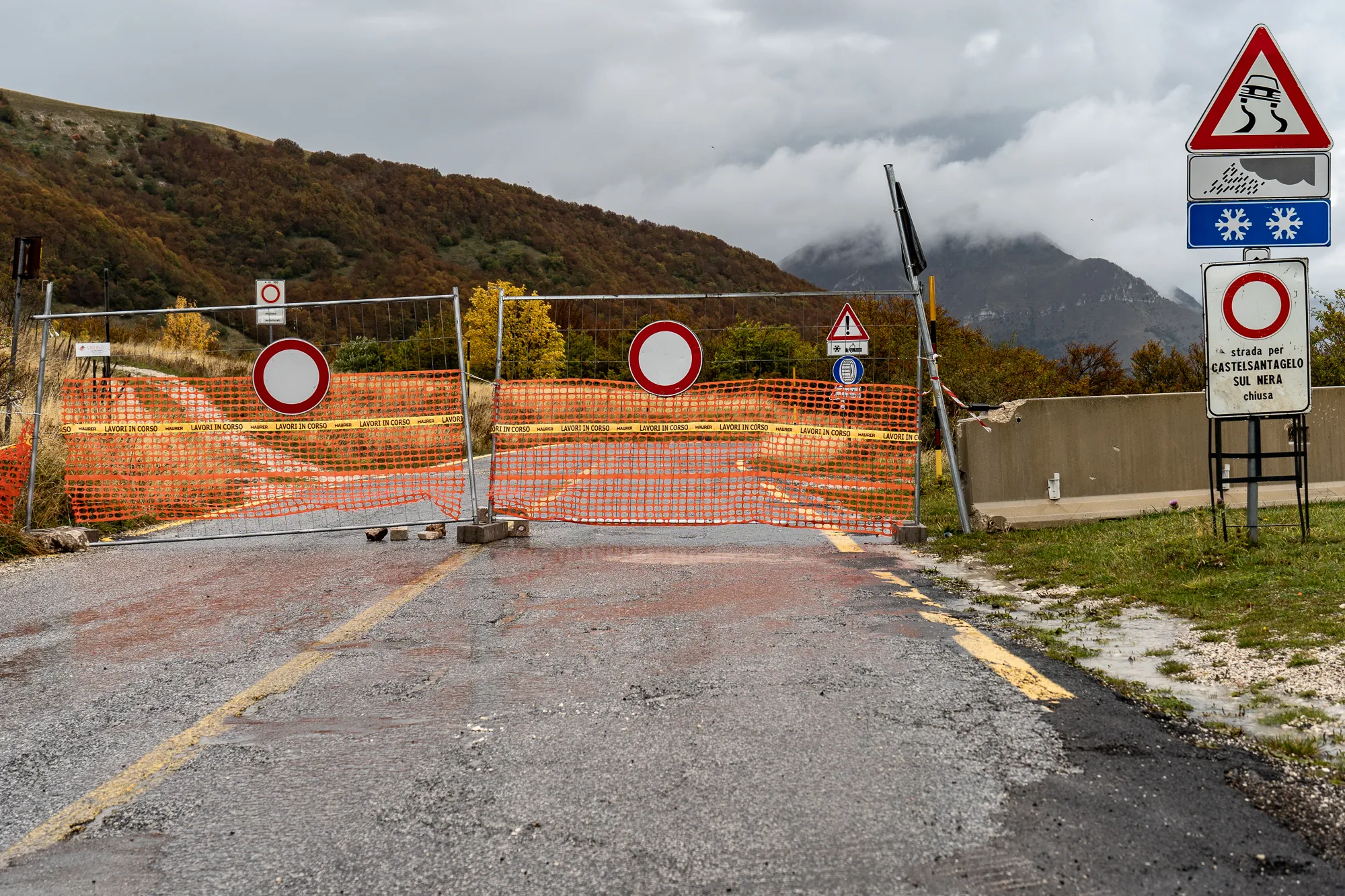 Gesperrte Straße mit orangefarbenem Absperrnetz und Verkehrsschildern im Bergland.