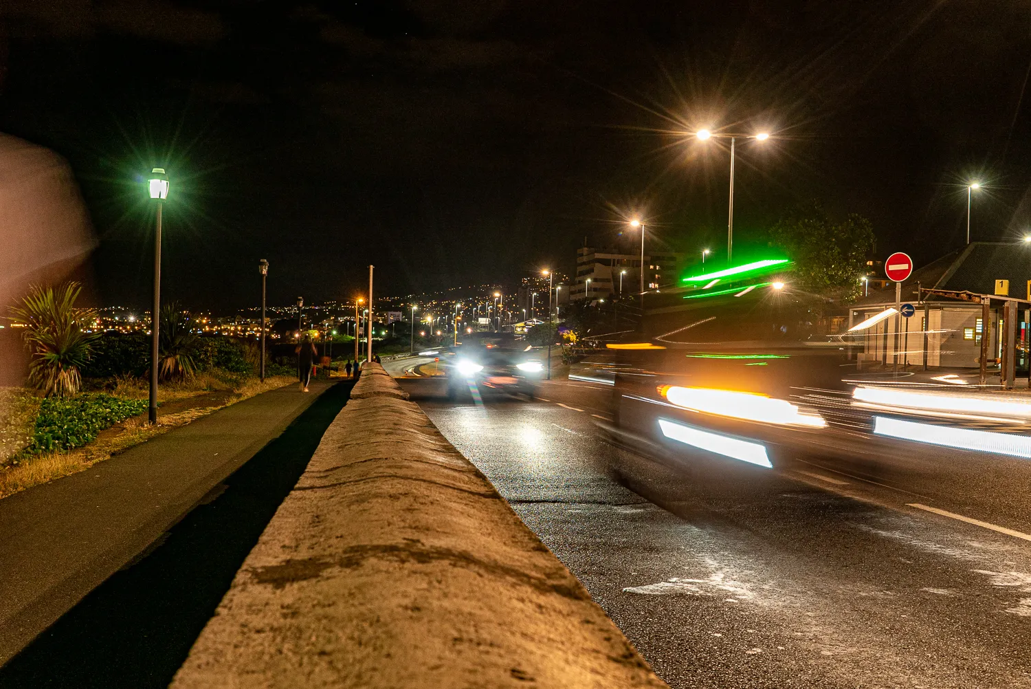 Nachtansicht einer beleuchteten Straße mit Lichtspuren von Autos und einem Gehweg.