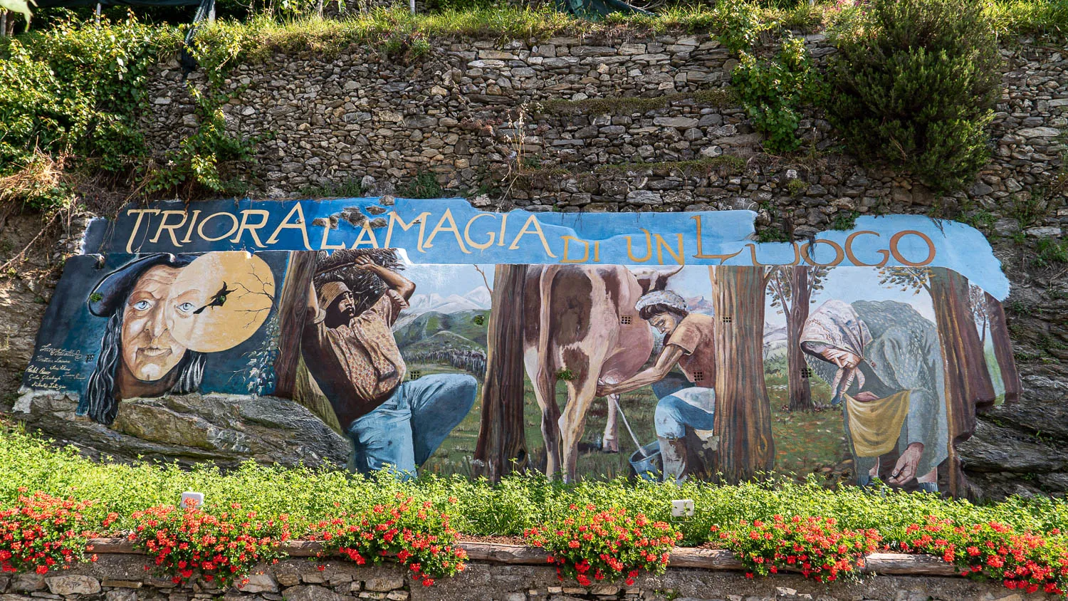Farbige Wandmalerei mit Figuren und Text vor Steinmauer und Blumenbeet
