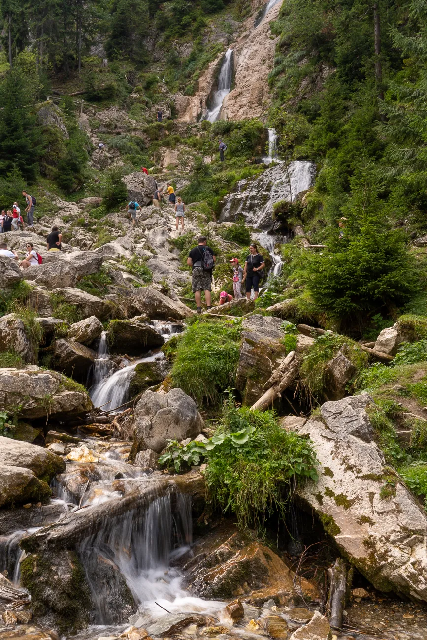 Wasserfall fließt über Felsen in einem bewaldeten Tal mit Wanderern