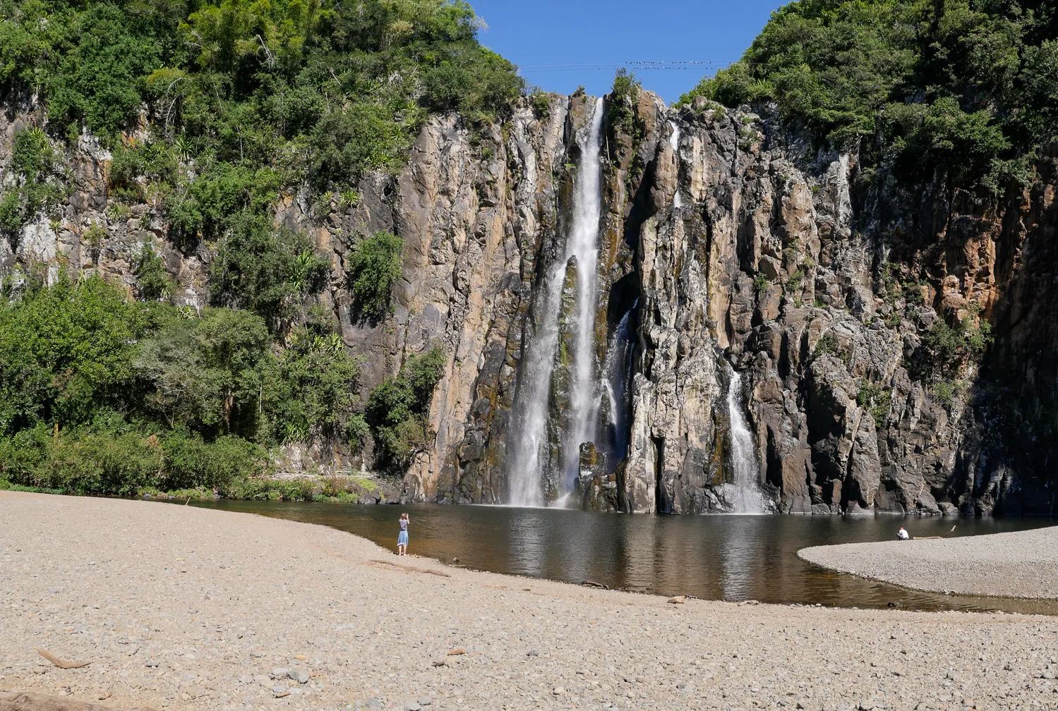 Wasserfall fällt von steilen Felsen in einen Teich mit Sandstrand.