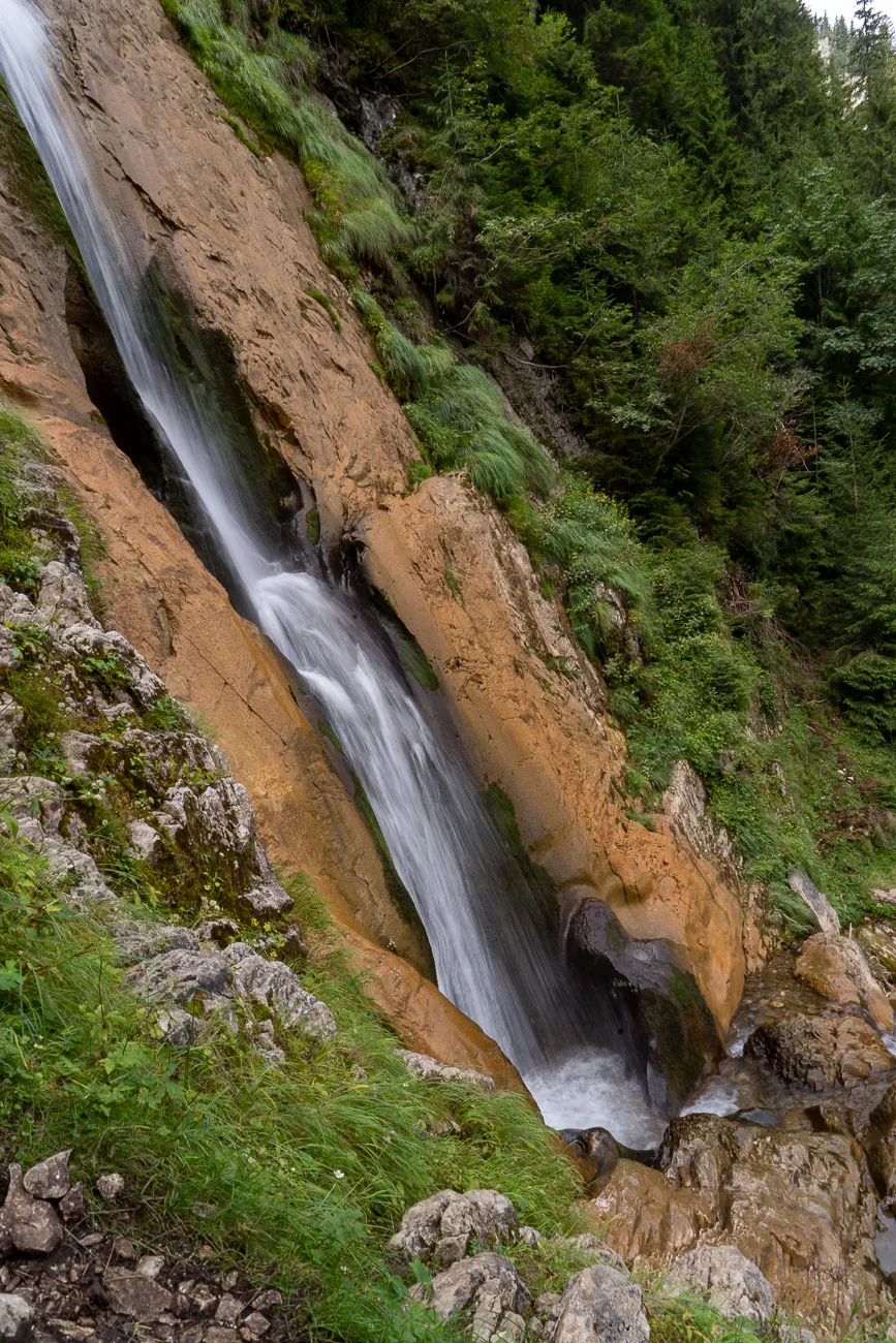 Wasserfall fließt über steinige, grüne Hangflanke in Wald.