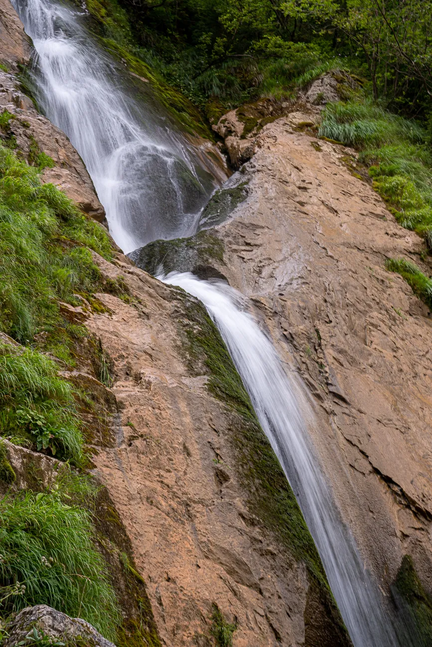 Wasserfall fließt über steinige Böschung mit grünem Moos und Gras