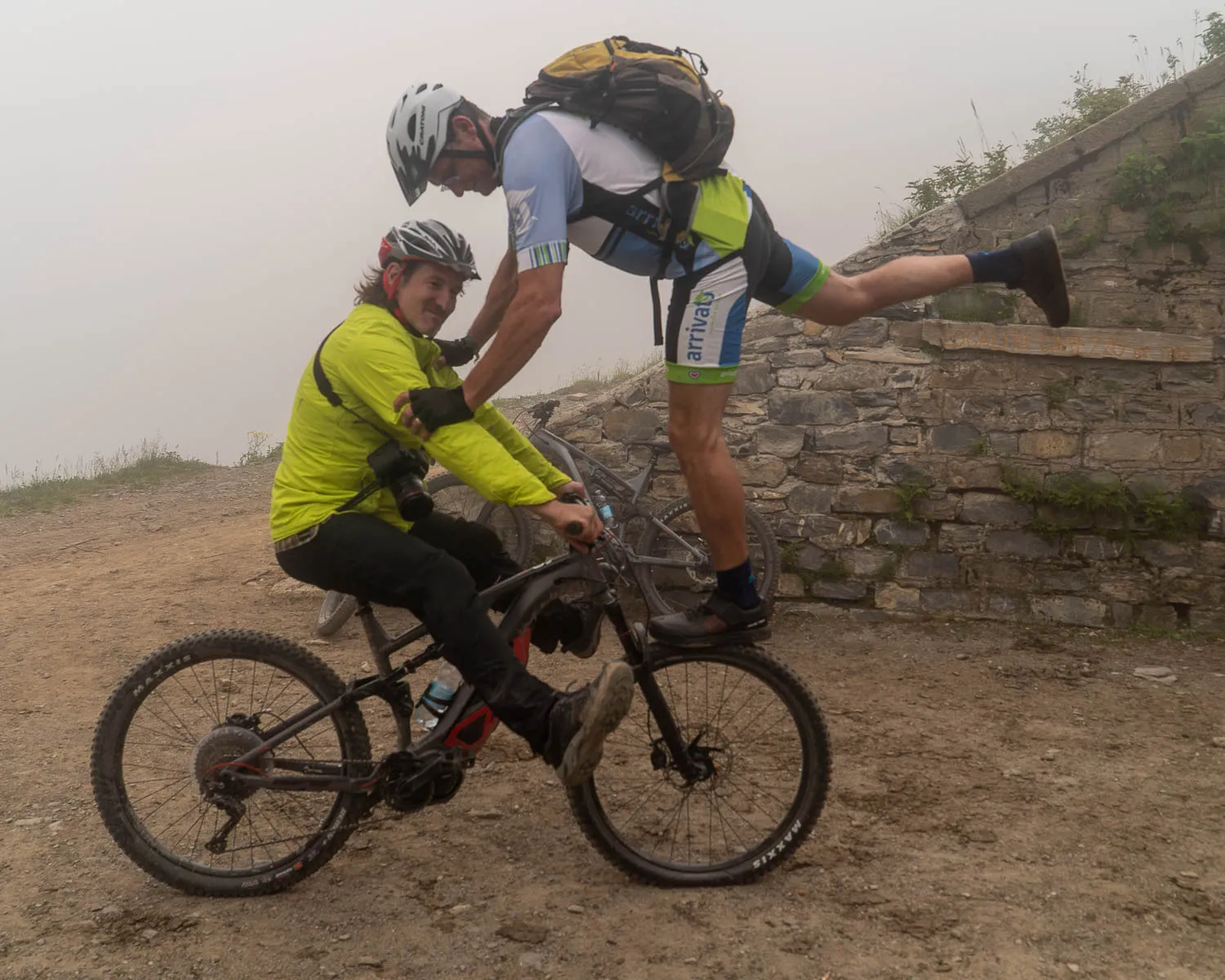 Zwei Radfahrer im Nebel, einer sitzt auf dem Fahrrad, der andere balanciert daneben.